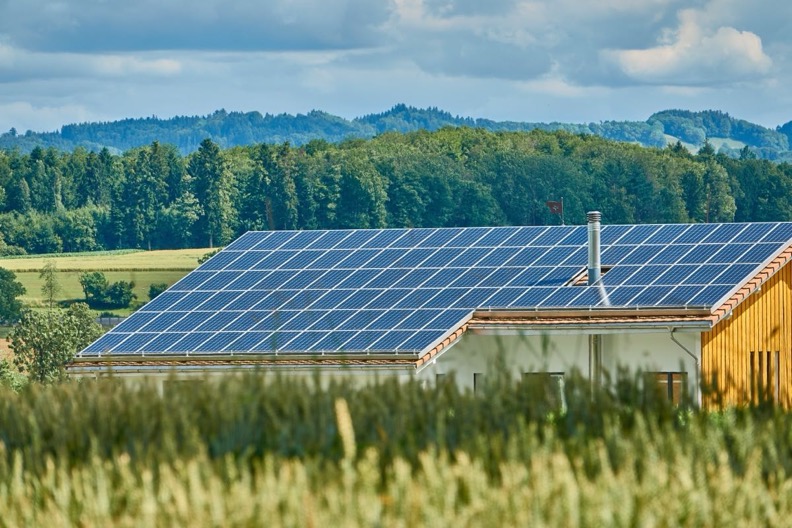 Cubierta de un edificio rural con toda su vertiente ocupada por módulos fotovoltaicos, campo de cereal en primer plano y bosque y colinas al fondo